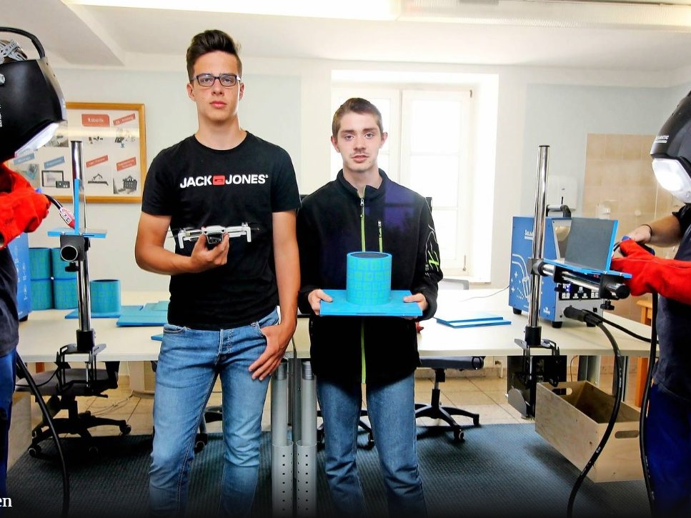 Schüler in einer Werkstatt: zwei Schweißer mit Helmen, einer hält eine Drohne, und einer präsentiert ein zylindrisches Modell.
