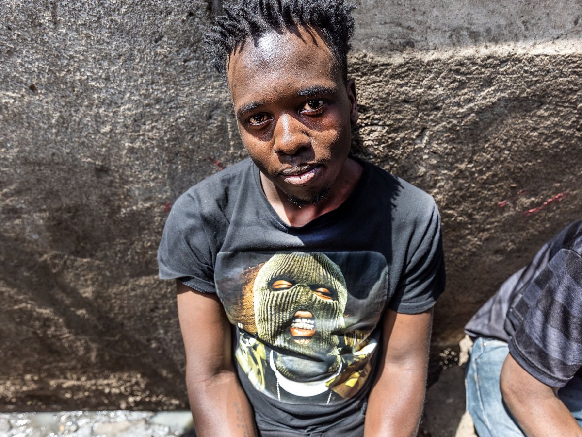 Porträt eines jungen Mannes mit Dreadlocks, der ein schwarzes T-Shirt mit Sturmhauben-Motiv trägt, vor einer strukturierten Wand.