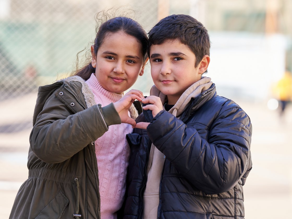 Zwei Kinder bilden mit ihren Händen ein Herz und lächeln in die Kamera, in Winterjacken gekleidet.