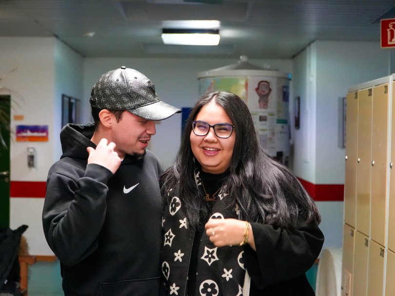 Zwei lächelnde Personen, ein Mann mit Kapuzenpullover und Kappe, und eine Frau mit Brille und gemustertem Schal, in einem Flur.