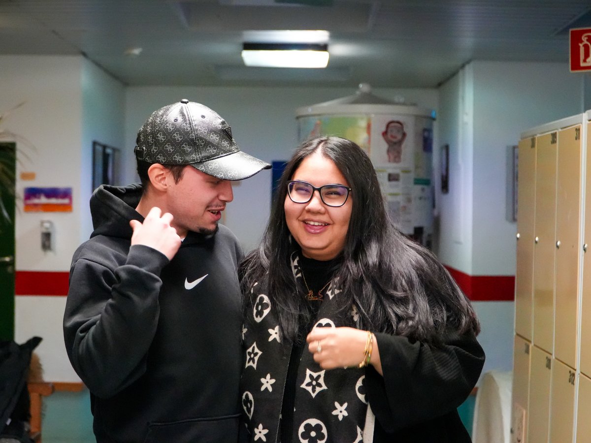 Zwei lächelnde Personen, ein Mann mit Kapuzenpullover und Kappe, und eine Frau mit Brille und gemustertem Schal, in einem Flur.