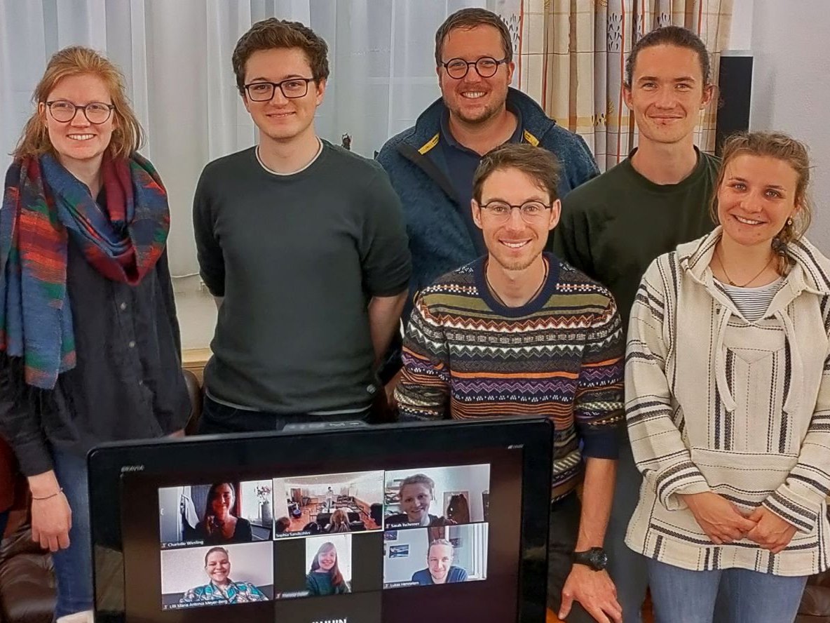 Eine lächelnde Gruppe junger Erwachsener in einem Raum, während ein Fernseher weitere Teilnehmer im Videoanruf zeigt.