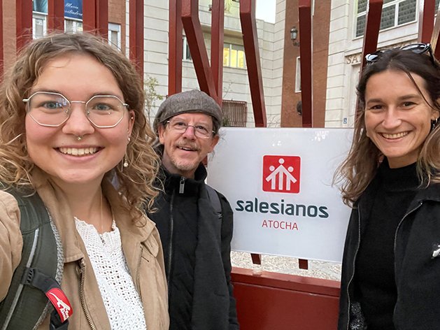 Drei lächelnde Personen, zwei Frauen und ein Mann, posieren vor einem Schild 'Salesianos ATOCHA'.