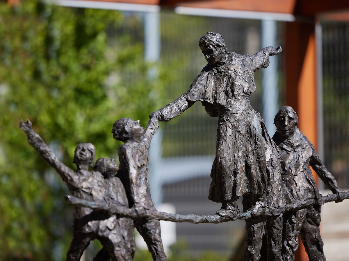 Texturierte Bronzeskulptur einer Gruppe von Menschen, die auf einem Balken balancieren, eine Frau greift nach einem Kind. Unscharfer grüner Hintergrund.