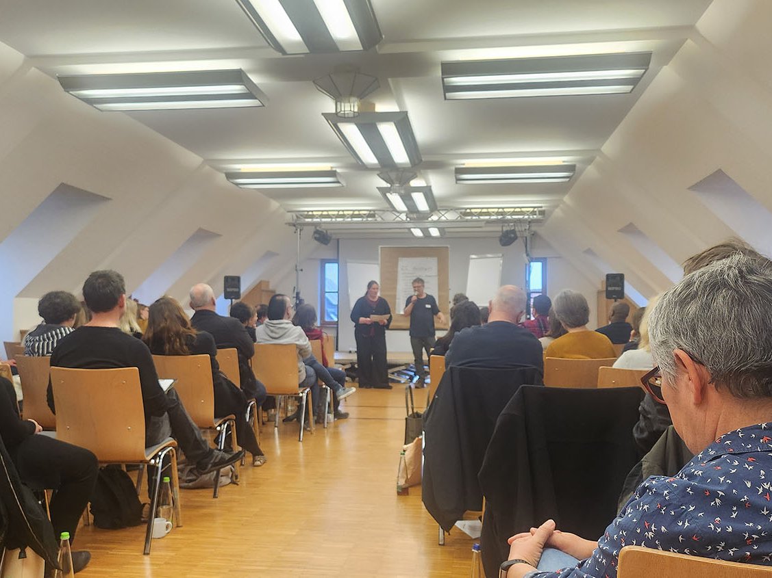 Ein Publikum sitzt zwei Vortragenden auf einer Bühne in einem hellen, modernen Dachsaal gegenüber.