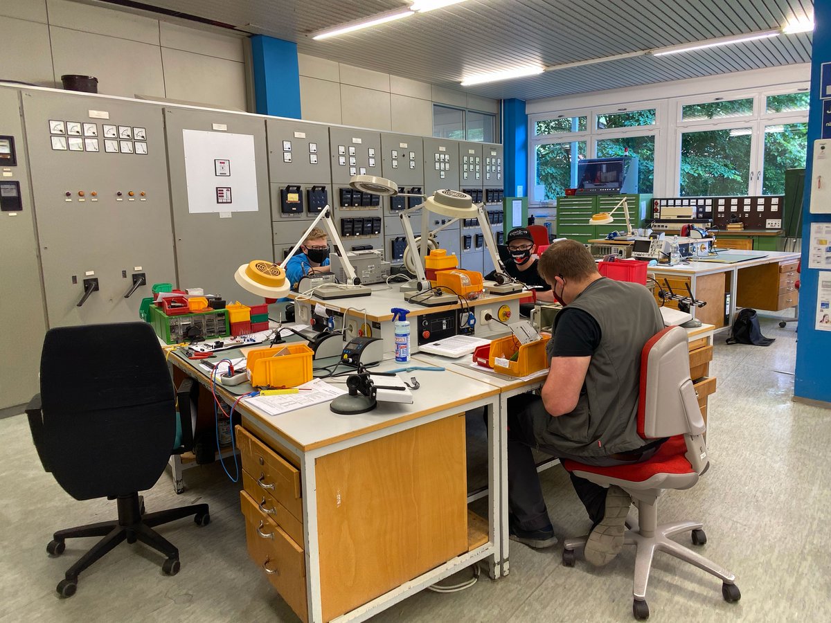 Drei Personen mit Masken arbeiten in einem Techniklabor an Tischen mit Elektronik und Messgeräten. Schaltpulte im Hintergrund.