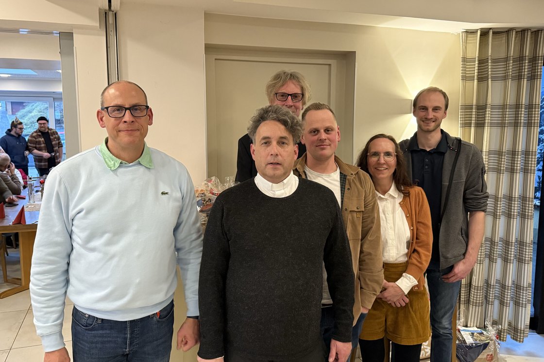 Eine Gruppe von sechs lächelnden Erwachsenen (fünf Männer, eine Frau) posiert für ein Foto bei einer Indoor-Veranstaltung.