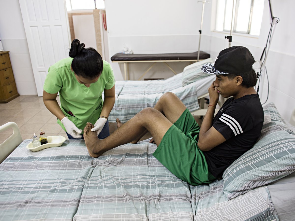 Eine Krankenschwester in grüner Kleidung versorgt den Fuß eines jungen Mannes, der auf einem Krankenhausbett sitzt.