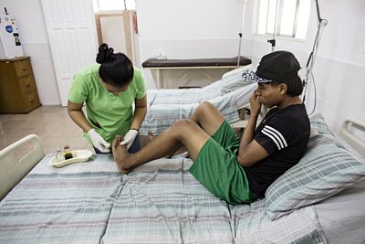 Eine Krankenschwester in grüner Kleidung versorgt den Fuß eines jungen Mannes, der auf einem Krankenhausbett sitzt.