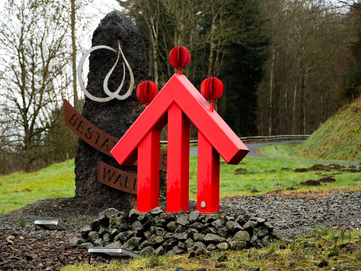 Rote pfeilförmige Skulptur mit drei Kugeln, Steinsäule mit "BESTA WALL"-Schild, auf steinigem, grasbewachsenem Hügel mit Bäumen.