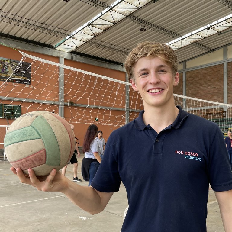 Lächelnder Don Bosco Freiwilliger hält einen abgenutzten Volleyball in einer Turnhalle mit Netz und Personen im Hintergrund.