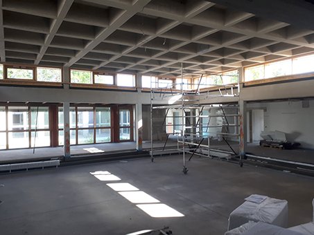 Geräumiger, leerer Raum in Renovierung mit markanter Gitterbetondecke, großen Fenstern und einem Rollgerüst.