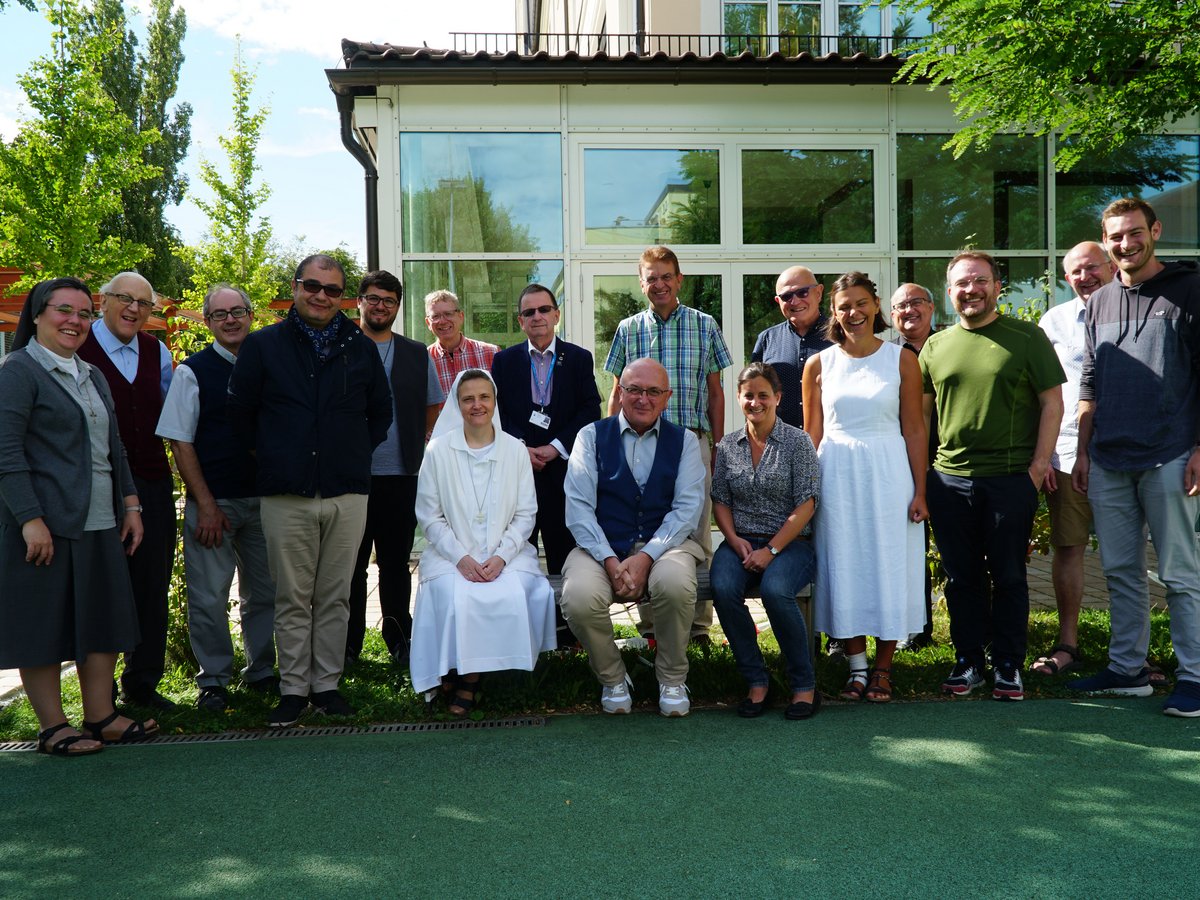 Gruppenfoto von 20 Personen, Männer und Frauen, inklusive zwei Nonnen, lächelnd vor einem modernen Gebäude mit großen Fenstern im Grünen.