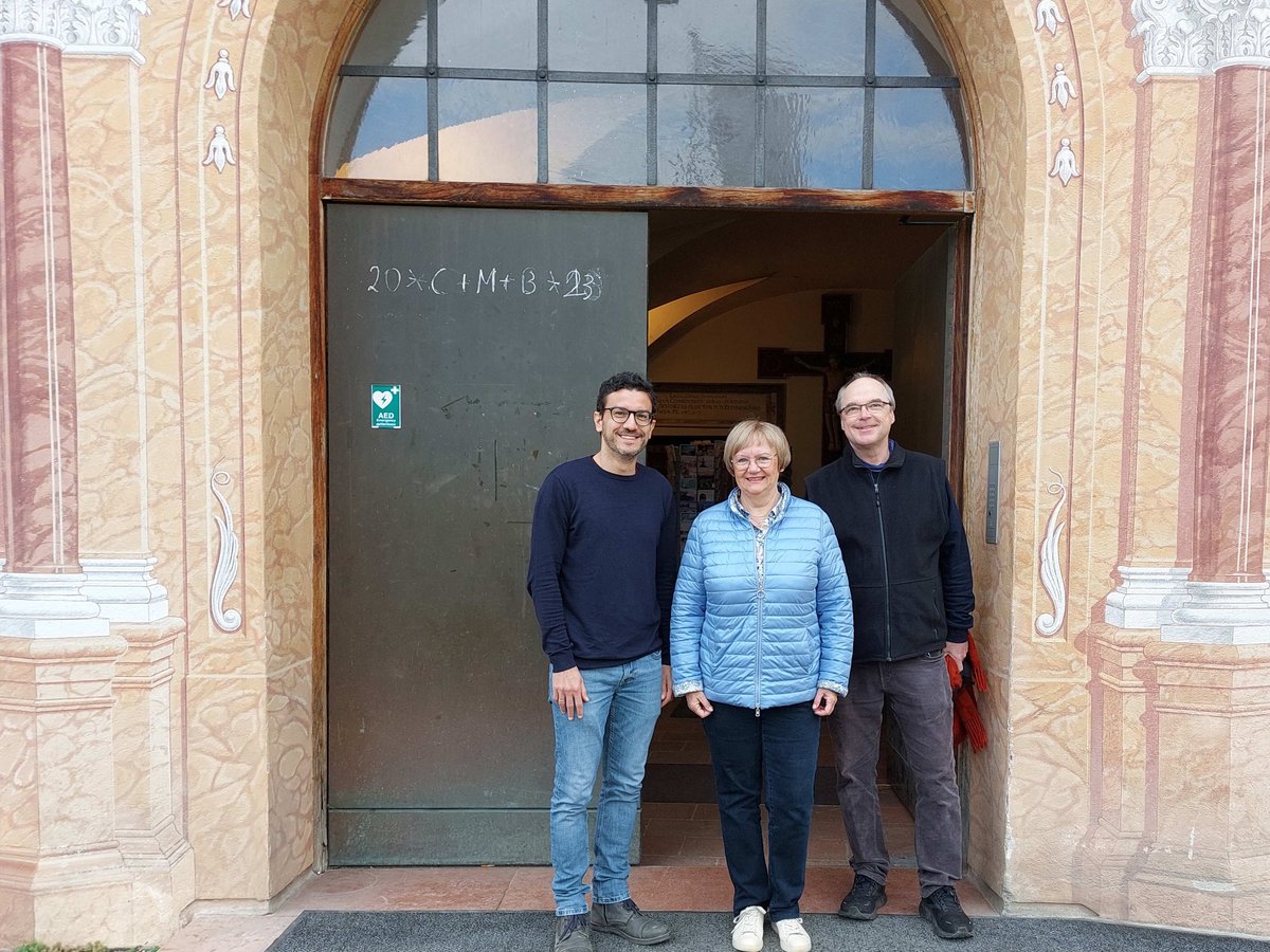 Drei Personen lächeln vor dem reich verzierten Eingang einer Kirche mit der Aufschrift '20*C+M+B*23'.