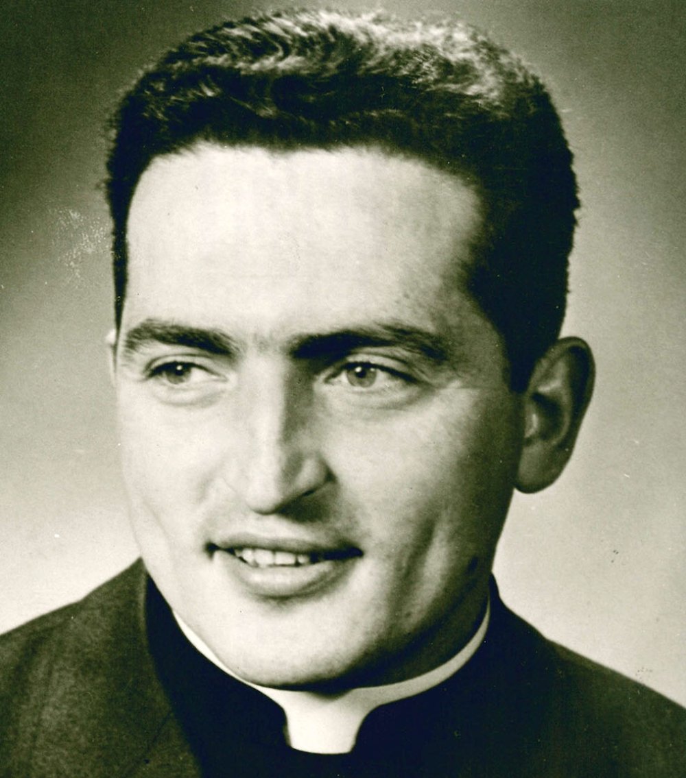 Halbporträt eines lächelnden jungen Priesters mit dunklem Haar und Kollar in Sepia-Tönen.