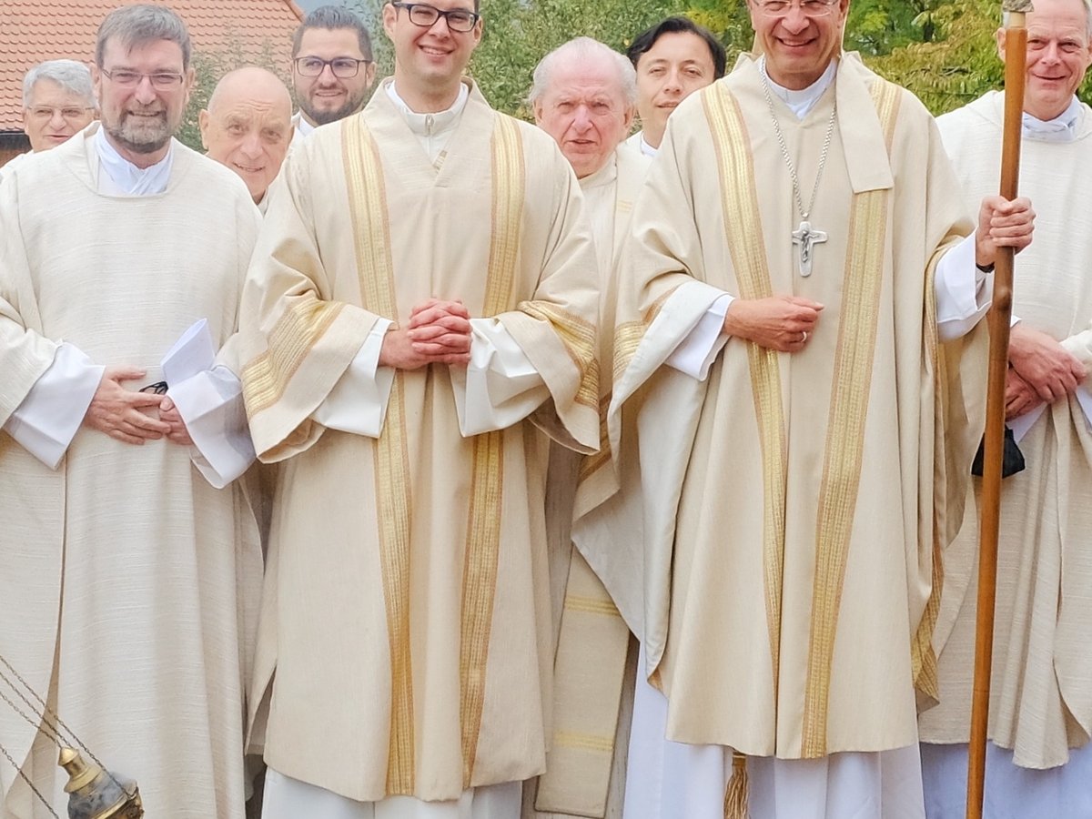 Eine Gruppe lachender katholischer Geistlicher in weißen Gewändern, darunter ein Bischof mit Mitra und Hirtenstab, im Freien.