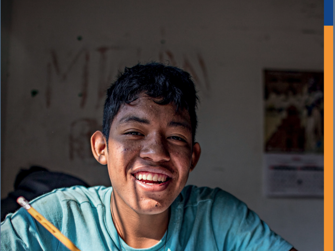 Ein lächelnder junger Mann in blauem Hemd schreibt an einem Tisch und blickt in die Kamera. Titelbild Don Bosco Mission Jahresbericht 2024.