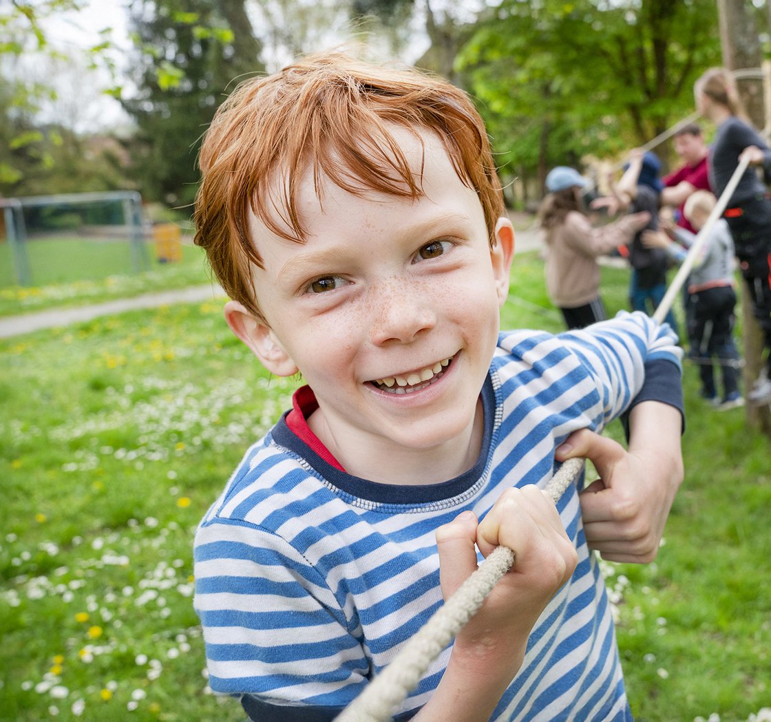 Lächelnder rothaariger Junge mit Sommersprossen hält ein Seil, spielt mit anderen Kindern bei einer Outdoor-Aktivität.