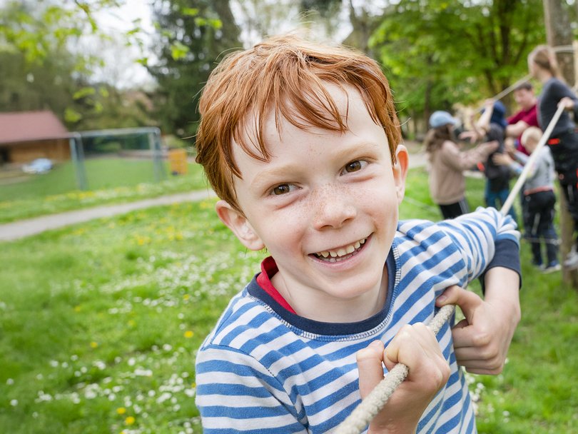 Lächelnder rothaariger Junge mit Sommersprossen hält ein Seil, spielt mit anderen Kindern bei einer Outdoor-Aktivität.