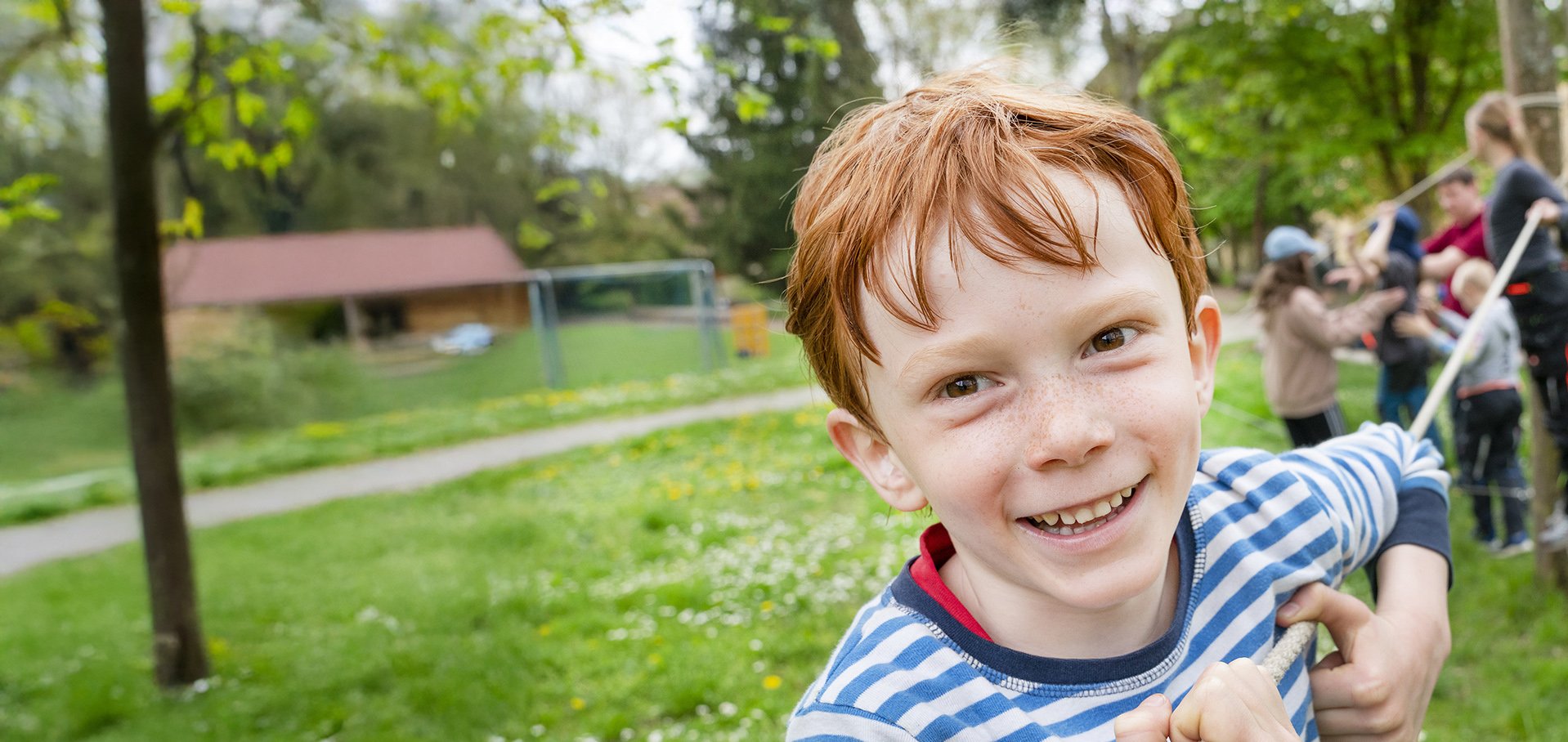 Lächelnder rothaariger Junge mit Sommersprossen hält ein Seil, spielt mit anderen Kindern bei einer Outdoor-Aktivität.
