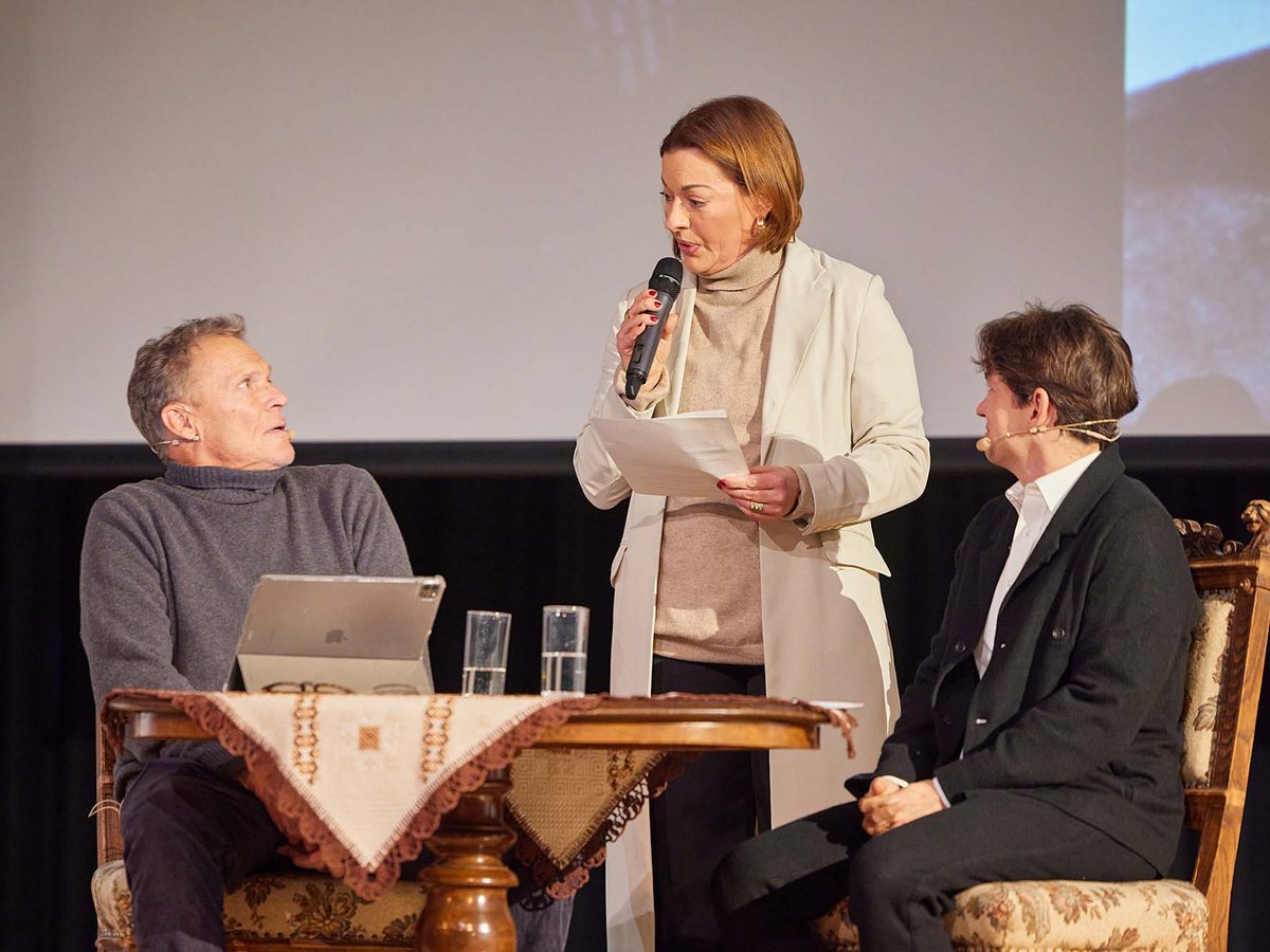 Drei Personen auf einer Bühne: eine Frau spricht ins Mikrofon, zwei Personen sitzen und hören zu bei einer Podiumsdiskussion.