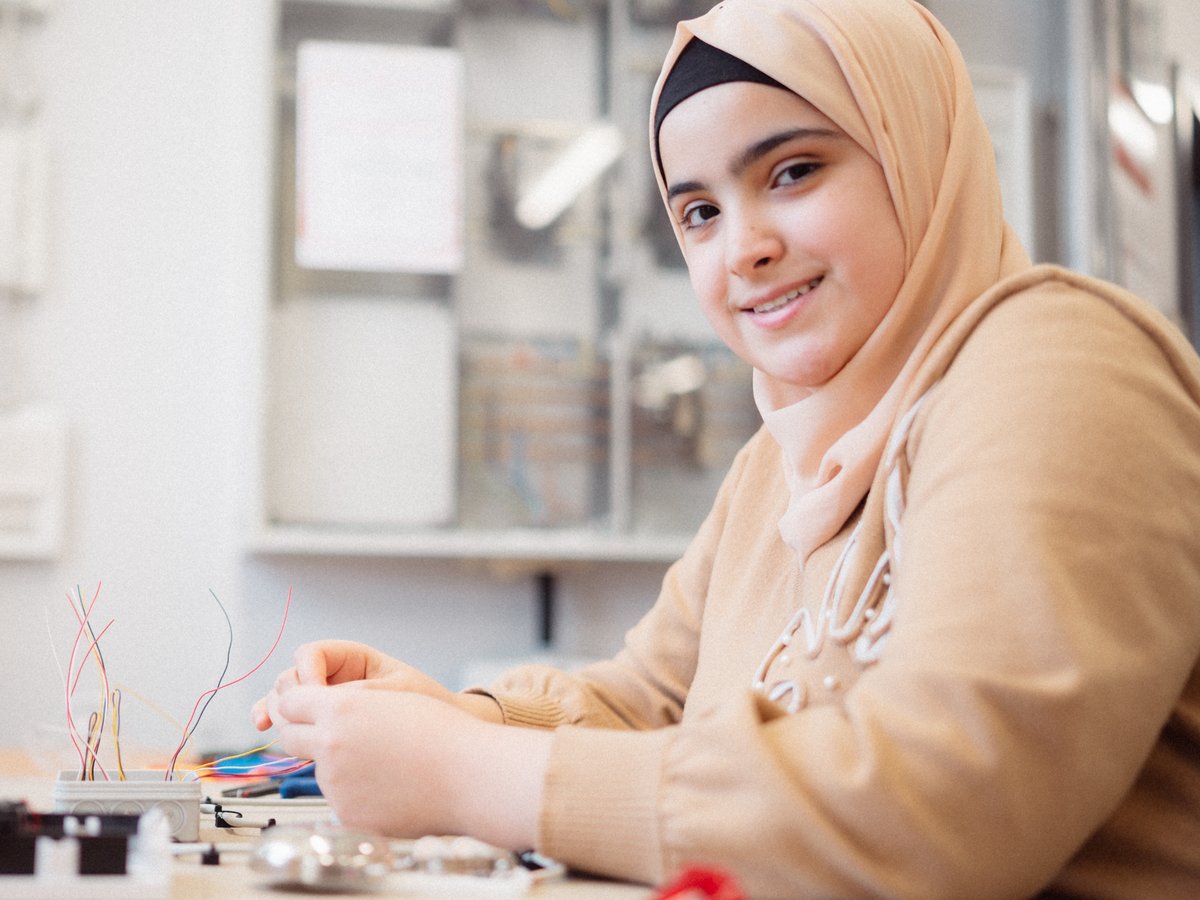 Eine lächelnde junge Frau mit beigem Hidschab arbeitet an einem Schreibtisch mit bunten Elektrokabeln und Werkzeugen.