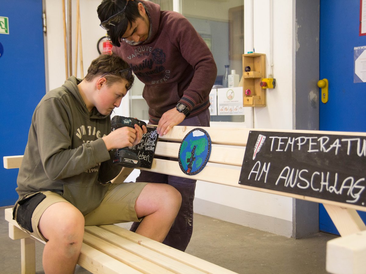 Zwei junge Männer bauen eine Holzbank mit Schildern zu Klimawandel und Erde zusammen; einer bohrt, der andere hilft.