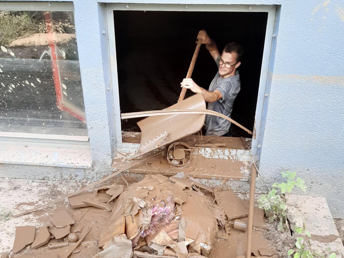 Mann mit Brille schaufelt dicken Schlamm aus einem Türrahmen in einem hochwassergeschädigten Gebäude, umgeben von Schlamm.