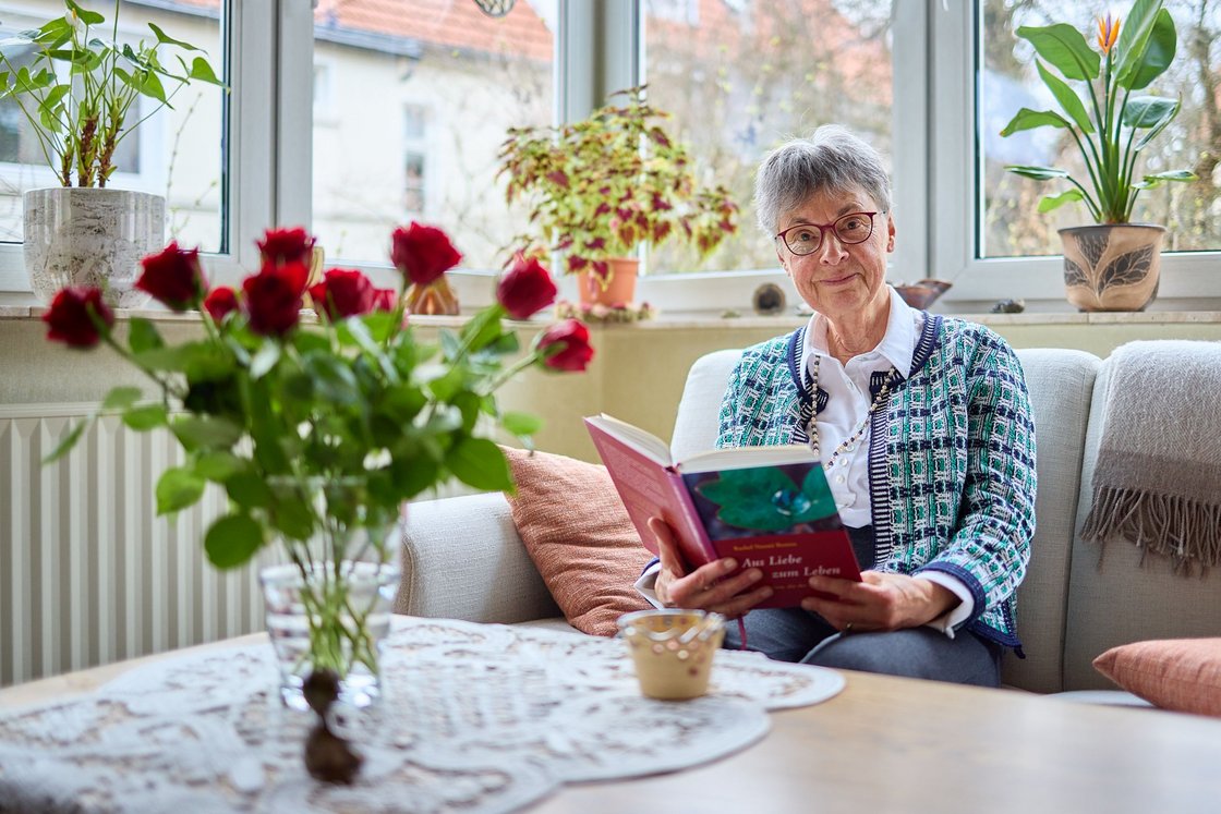 ältere Dame mit Kurzhaarfrisur und roter Brille sitzt lächelnd auf dem Sofa mit einem Buch, davor ein Strauß roter Rosen 