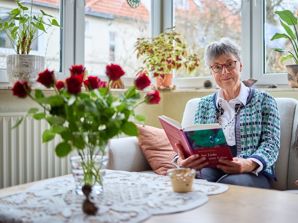 Elisabeth Middendorf ältere Dame mit Kurzhaarfrisur und roter Brille sitzt lächelnd auf dem Sofa mit einem Buch, davor ein Strauß roter Rosen