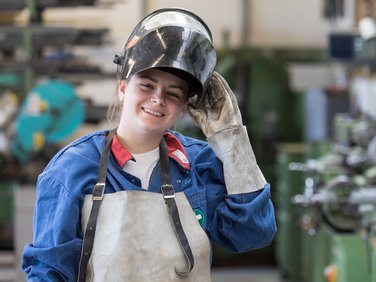 Lachende junge Auszubildende in Schutzkleidung hält ihre Schweißmaske hoch, im Hintergrund eine Werkstatt.