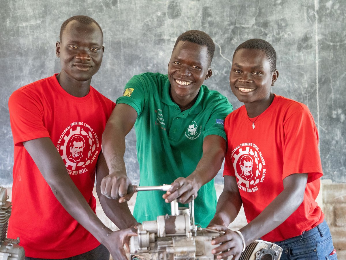 Drei lächelnde junge Männer in einer Berufsausbildungswerkstatt, die gemeinsam an einem Motor arbeiten.