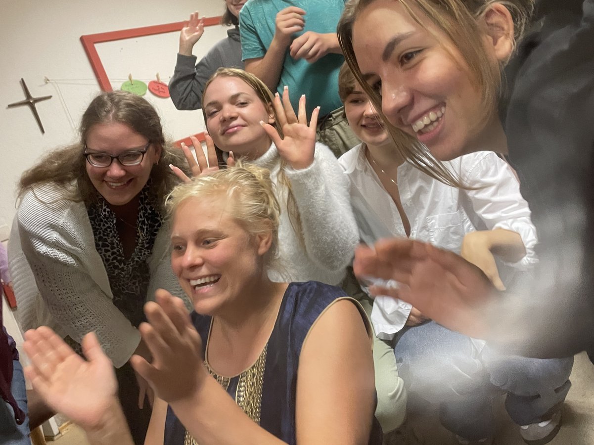 Eine Gruppe fröhlicher Jugendlicher lächelt und winkt in die Kamera in einem beleuchteten Raum.