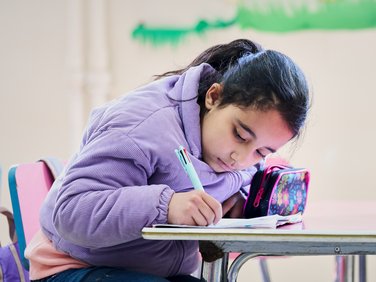 Ein konzentriertes junges Mädchen mit dunklen Haaren und lila Kapuzenpullover schreibt mit einem bunten Stift in ein Notizbuch an einem Schreibtisch.