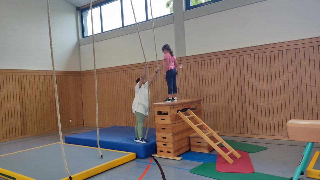 Kind steht in Sporthalle von Don Bosco Pfaffendorf auf Kasten und wird von Betreuerin unterstützt