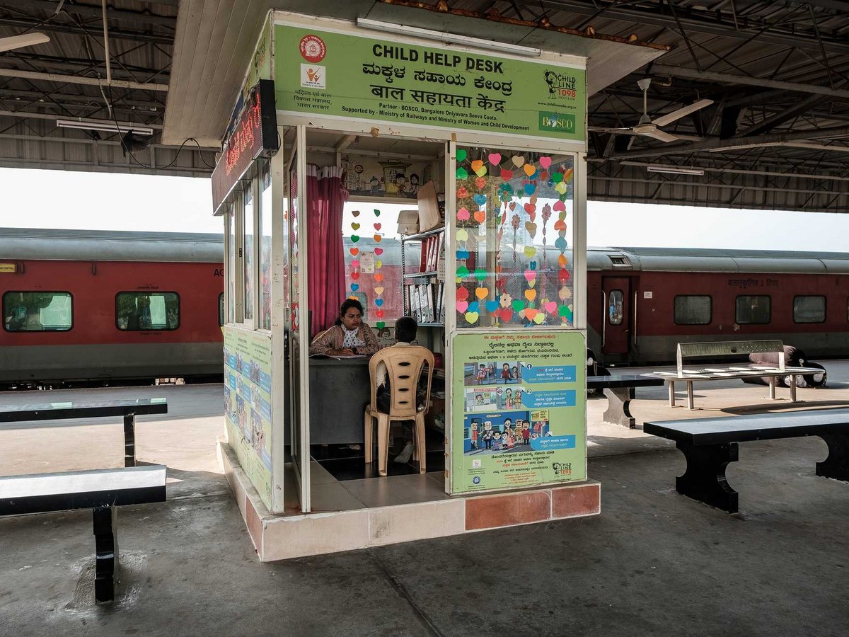 Bunte Kinderhilfsstelle an einem Bahnhof, eine Frau hilft einer Person, ein roter Zug im Hintergrund.