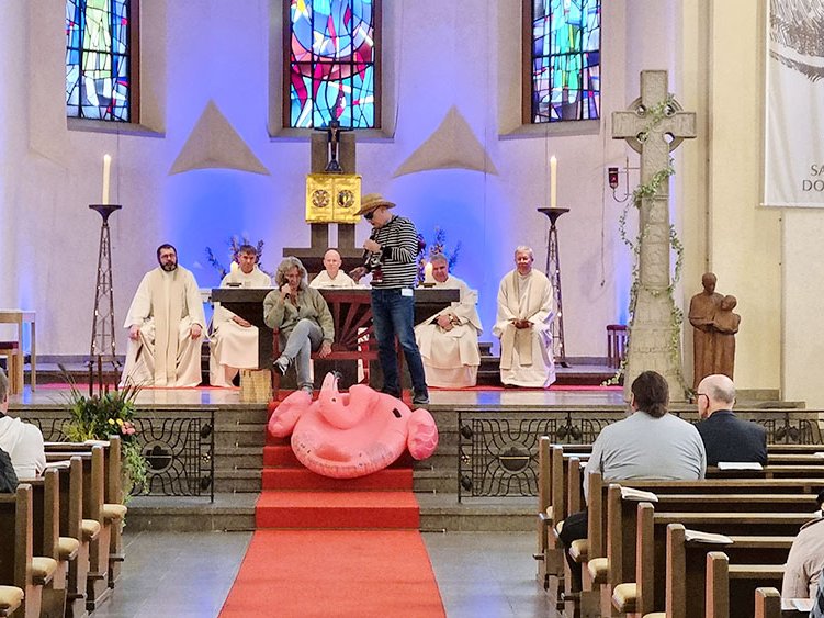 Ungewöhnlicher Gottesdienst in einer Kirche mit Redner im Strohhut, einer Frau und einem rosa Flamingo auf den Altarstufen.
