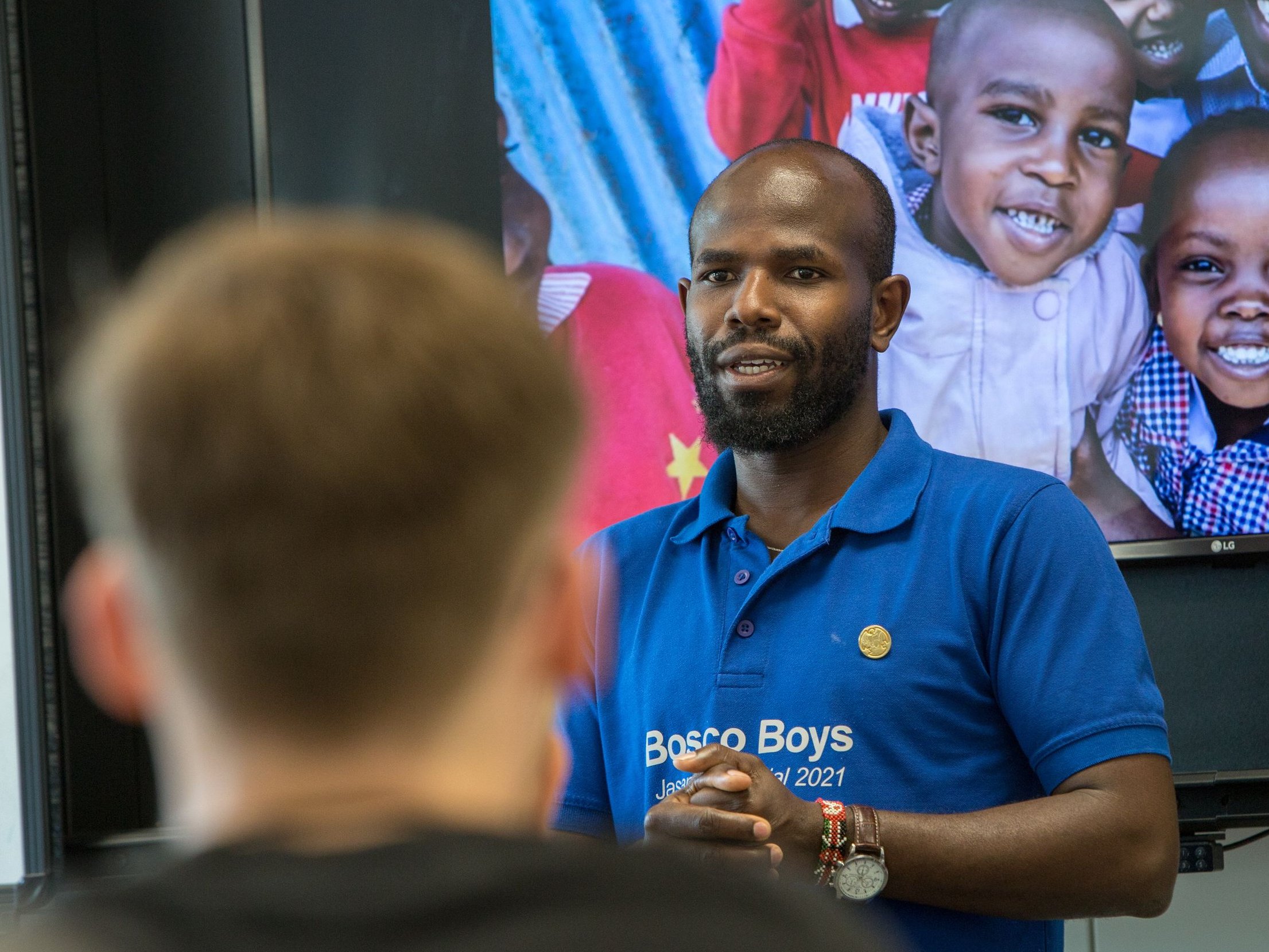 Ein Mann im 'Bosco Boys' Hemd spricht; dahinter ein Bildschirm mit lächelnden afrikanischen Kindern und einem Erwachsenen.