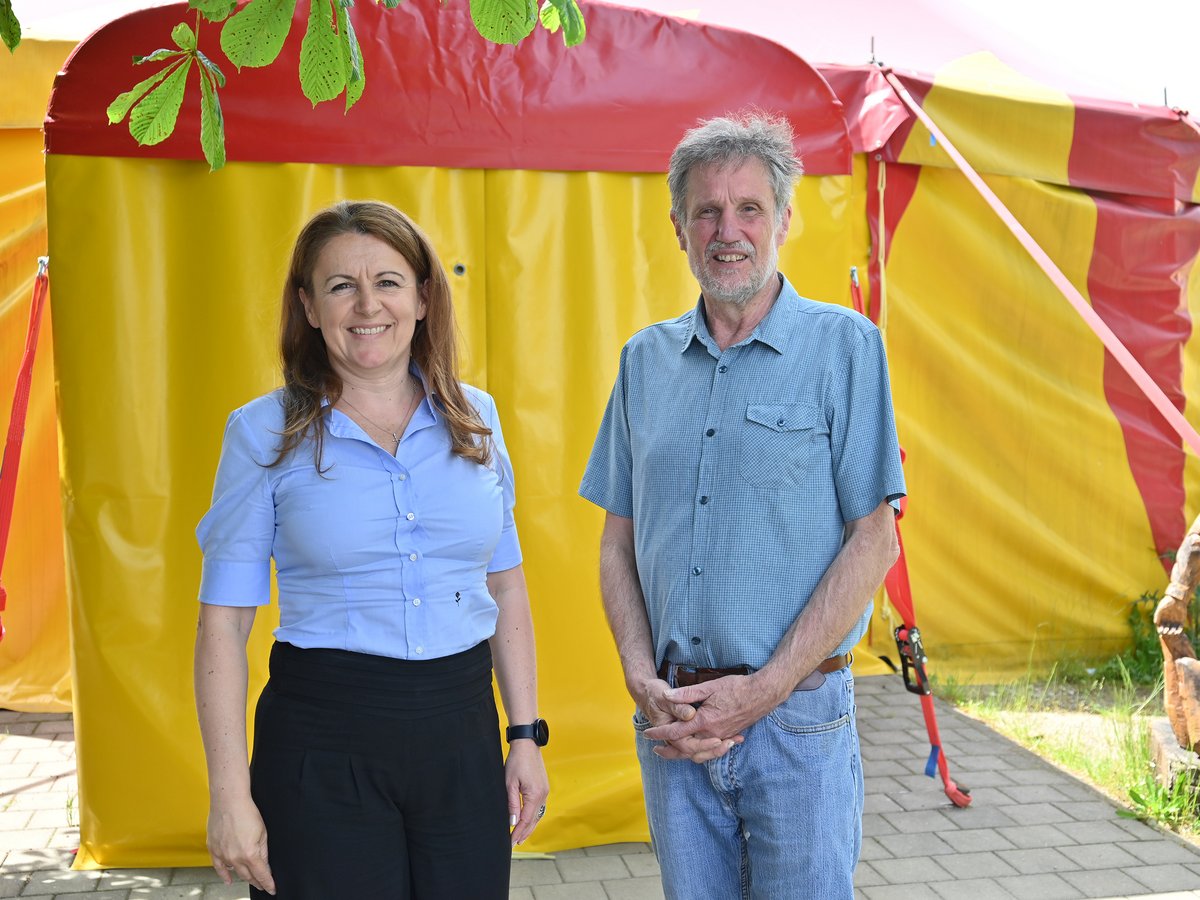 Eine lächelnde Frau und ein Mann stehen vor einem gelb-roten Zelt, mit grünen Blättern im Vordergrund.
