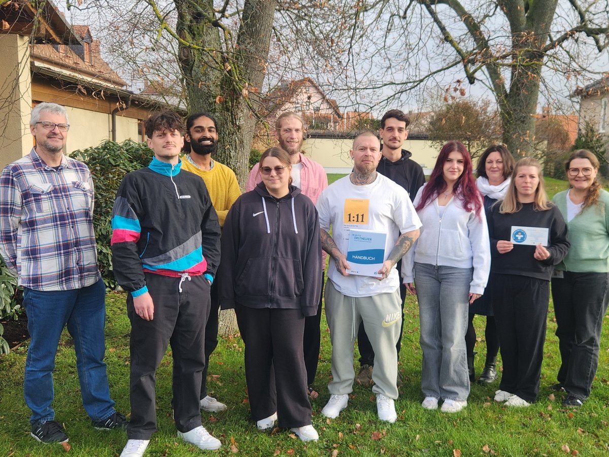 Diverse Gruppe von zwölf lächelnden Erwachsenen im Freien, ein Mann hält ein Erste-Hilfe-Handbuch, eine Frau ein Zertifikat.