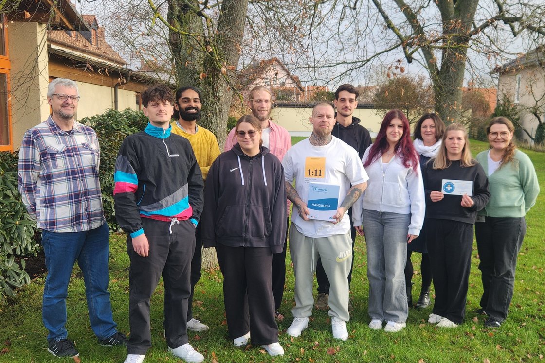 Diverse Gruppe von zwölf lächelnden Erwachsenen im Freien, ein Mann hält ein Erste-Hilfe-Handbuch, eine Frau ein Zertifikat.