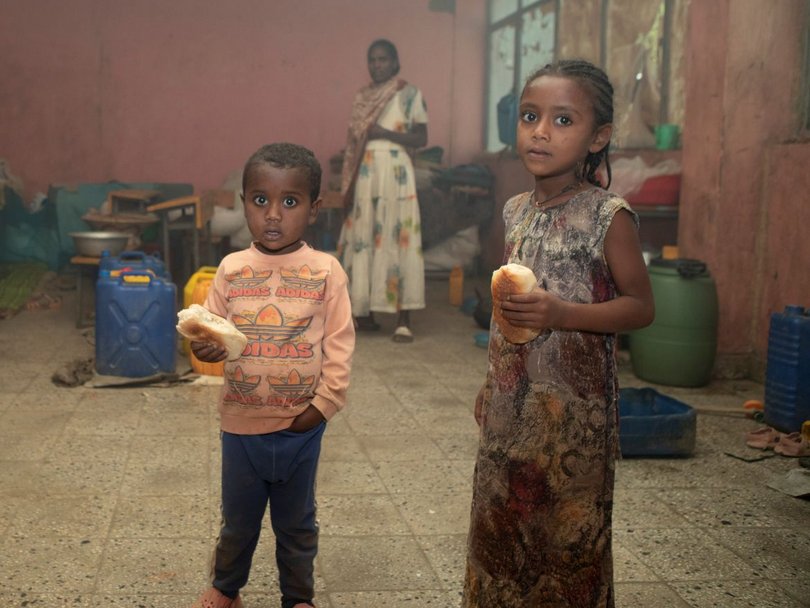 Zwei Kinder halten Brot und blicken in die Kamera, in einem überfüllten Raum, wahrscheinlich einer Unterkunft. Erwachsene sind im Hintergrund zu sehen.