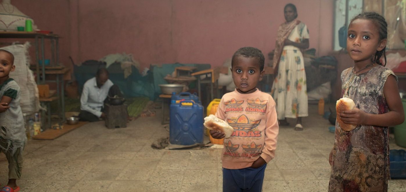 Zwei Kinder halten Brot und blicken in die Kamera, in einem überfüllten Raum, wahrscheinlich einer Unterkunft. Erwachsene sind im Hintergrund zu sehen.
