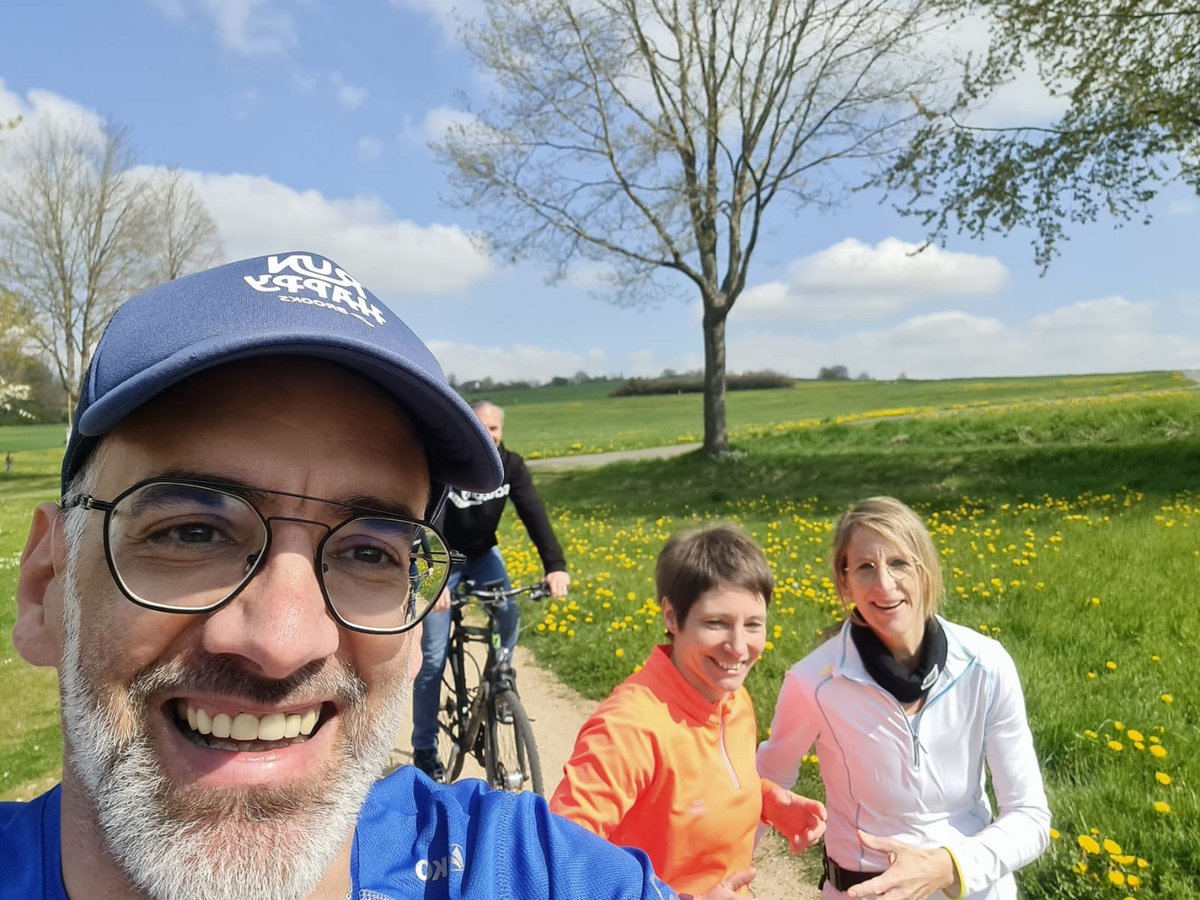 Lächelnder Mann macht ein Selfie mit Freunden beim Radfahren und Laufen in einer sonnigen Löwenzahnwiese.