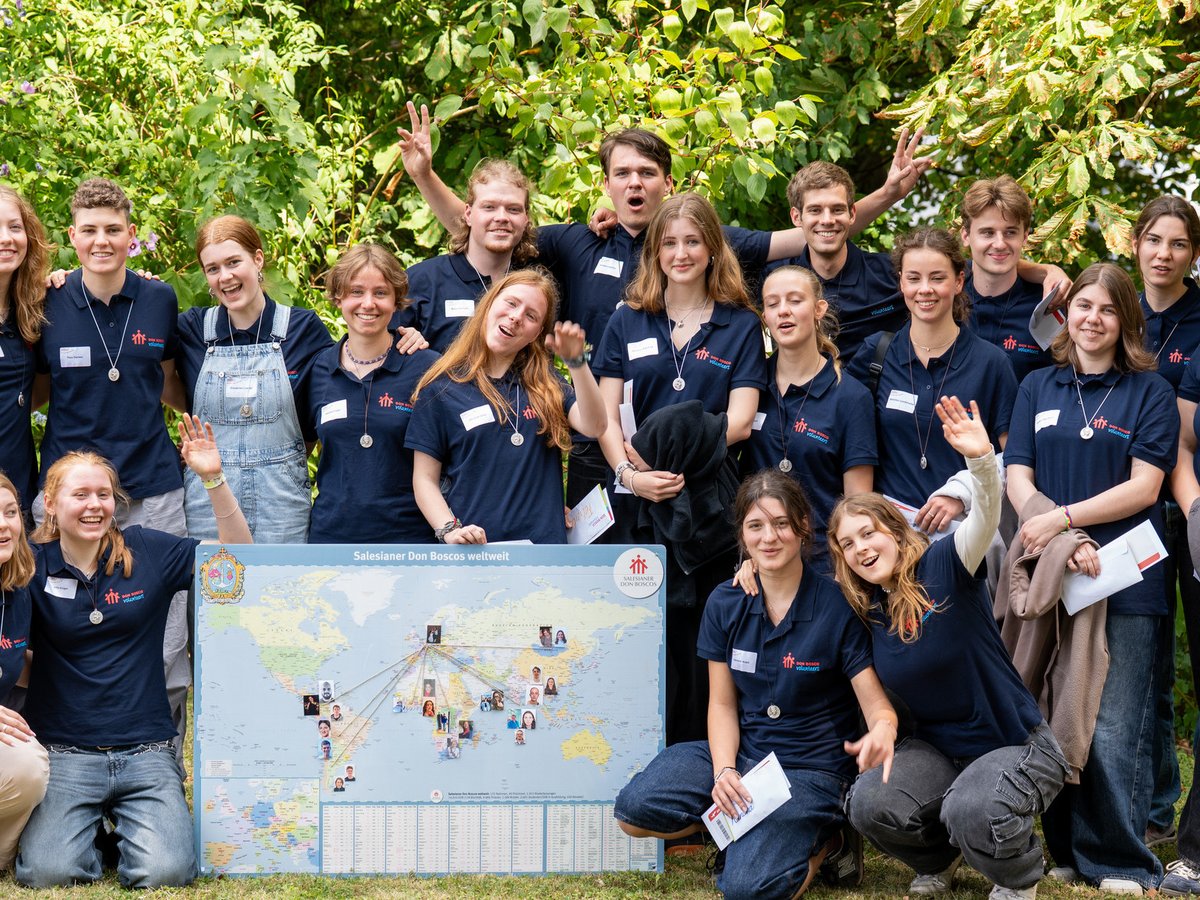 Eine Gruppe lächelnder junger Don Bosco Freiwilliger in blauen Shirts und Medaillons posiert mit einer Weltkarte draußen.