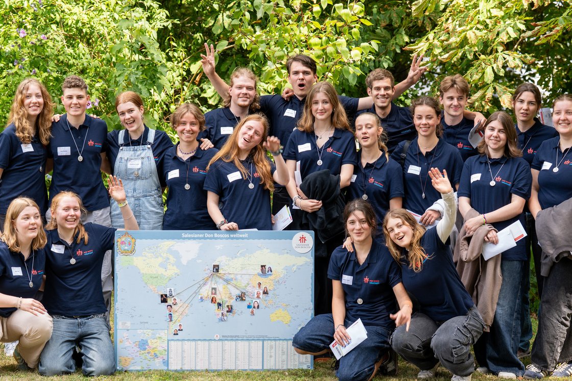 Eine Gruppe lächelnder junger Don Bosco Freiwilliger in blauen Shirts und Medaillons posiert mit einer Weltkarte draußen.