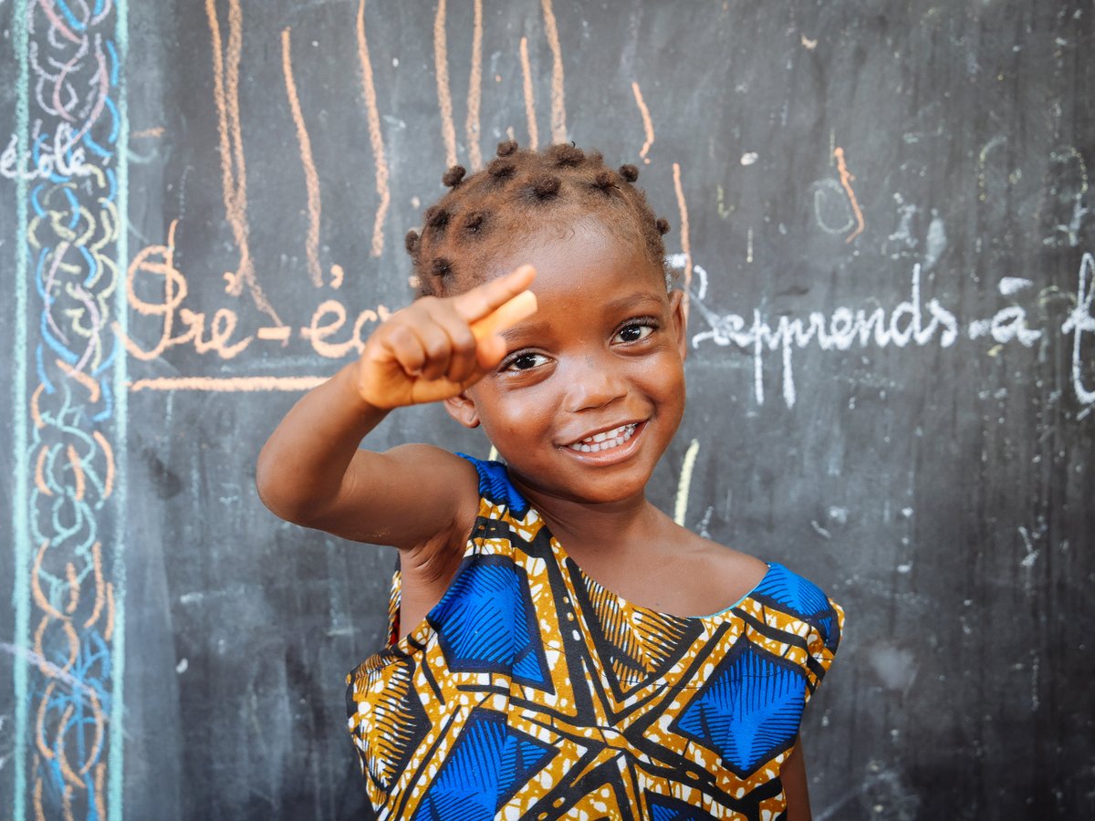 Ein lächelndes Mädchen mit Zöpfen zeigt auf die Kamera, vor einer Tafel mit Schriftzügen und bunten Zeichnungen.