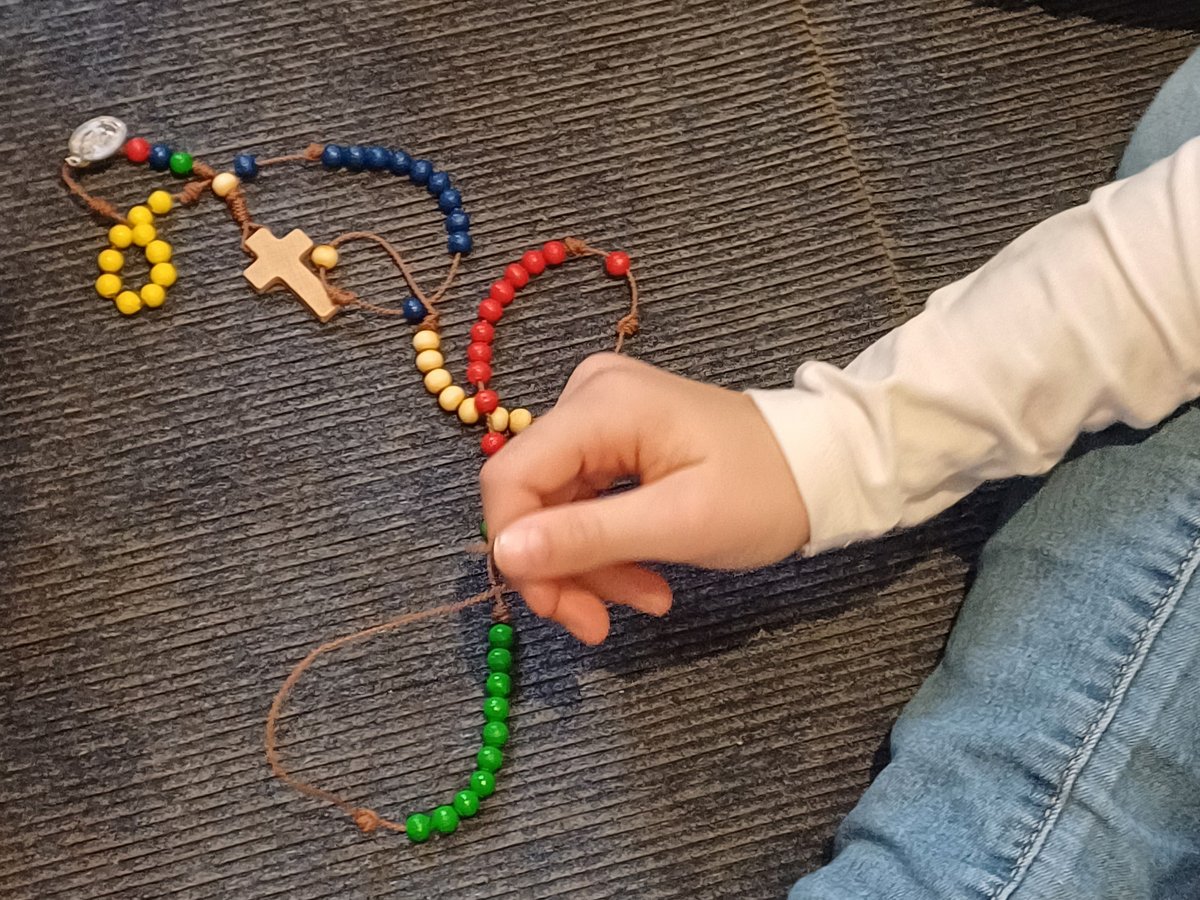 Kinderhand hält einen farbenfrohen Rosenkranz mit Holzperlen, einem Kreuz und einer Medaille auf dunklem Untergrund.