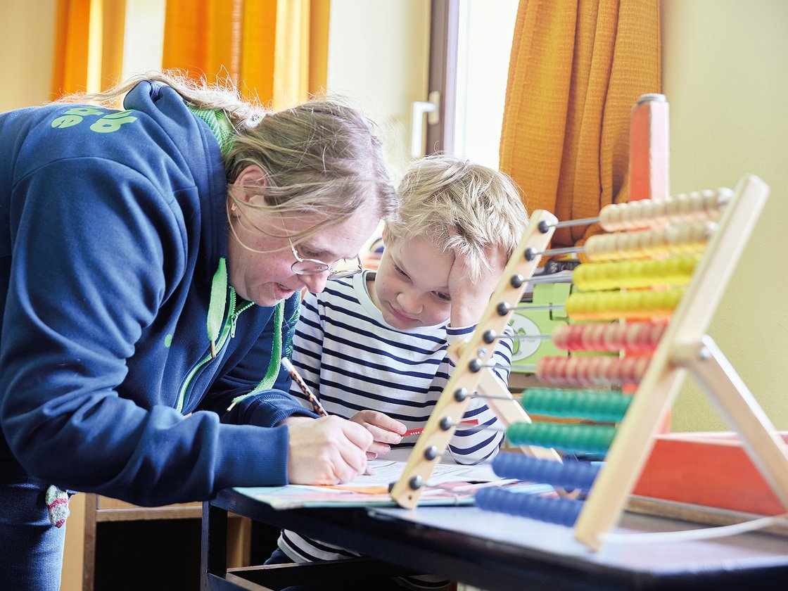 Eine Frau hilft einem kleinen Jungen bei den Schularbeiten an einem Tisch. Ein Abakus mit bunten Perlen ist im Vordergrund.