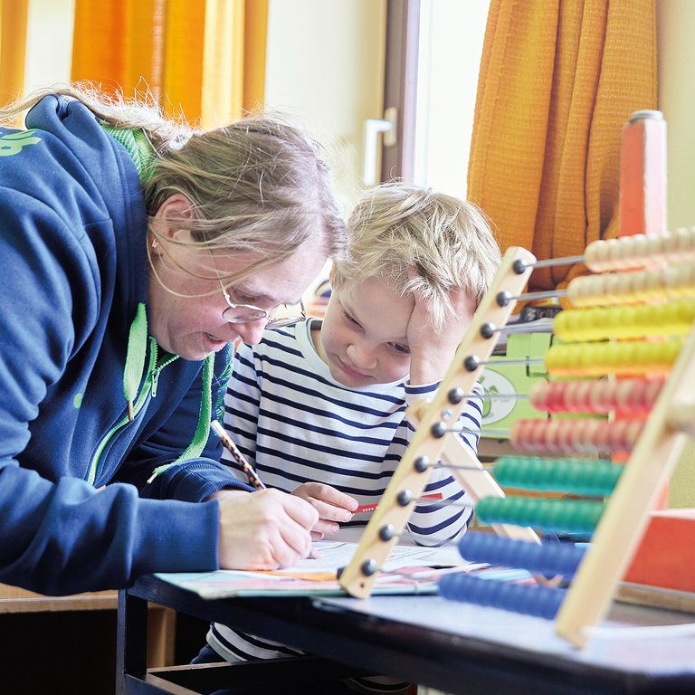 Eine Frau hilft einem kleinen Jungen bei den Schularbeiten an einem Tisch. Ein Abakus mit bunten Perlen ist im Vordergrund.